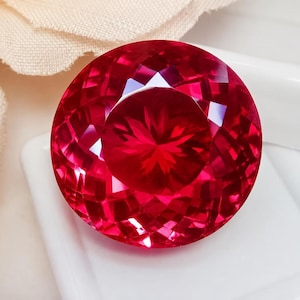 May include: A round, faceted gemstone with a deep, vibrant red color. The stone features a complex, geometric cut with numerous triangular and diamond-shaped facets that create a brilliant, sparkling effect across its entire surface.