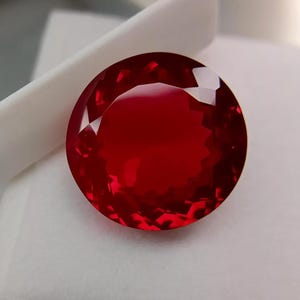 Pode incluir: Uma pedra preciosa redonda e facetada de cor vermelha profunda e vibrante. A pedra apresenta um corte brilhante com numerosas facetas triangulares e em forma de pipa que refletem a luz, criando uma aparência clara e translúcida.