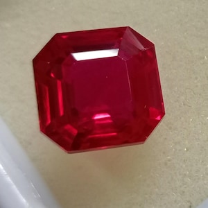 May include: A vibrant, deep red gemstone with an octagonal step-cut shape. The stone features sharp, geometric facets that create a symmetrical pattern, reflecting light with high clarity and a rich, saturated crimson hue throughout the surface.