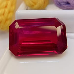 Peut inclure: Une pierre précieuse rouge vif et profonde taillée en forme d'émeraude rectangulaire. La pierre présente une surface à facettes qui reflète la lumière, créant des reflets et des ombres internes clairs sur son corps cramoisi transparent.