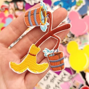 May include: Embroidered patch featuring a yellow broom and two orange buckets with blue accents. The patch is held in a hand, surrounded by other colorful patches with cartoon characters.
