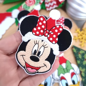 Peut inclure: Écussons brodés représentant Minnie Mouse avec un chapeau de Père Noël, un nœud rouge à pois blancs. Autres écussons : chapeau de Père Noël, nœuds et écusson Daisy Duck. Décorations de Noël.