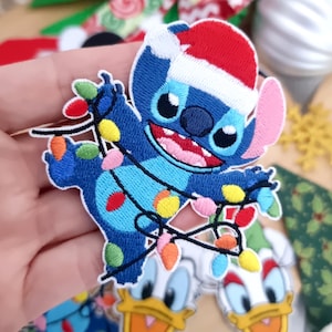 Op de afbeelding: Geborduurde patch met het personage Stitch uit Lilo & Stitch, met een kerstmuts en kerstverlichting. De patch is blauw, met rode, gele en roze accenten. Andere patches met Donald Duck en kerstversieringen staan op de achtergrond.
