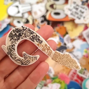 May include: Embroidered patch in the shape of a pirate hook, featuring intricate beige and black detailing. The hook is held against a backdrop of various colorful patches, showcasing a craft or hobby theme.