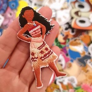 Könnte beinhalten: Gestickter Aufnäher mit einer Zeichentrickfigur mit langen schwarzen Haaren, die ein rot-weißes Oberteil und einen gemusterten Rock trägt. Der Aufnäher wird in einer Hand gehalten, mit einem Hintergrund aus anderen bunten Aufnähern.