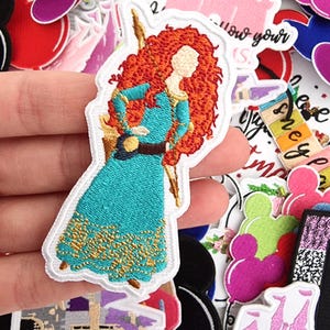 Może przedstawiać: Haftowana naszywka przedstawiająca postać z kreskówki, kobietę z długimi, rudymi kręconymi włosami, ubraną w turkusową suknię i trzymającą laskę. Naszywka otoczona jest innymi kolorowymi naszywkami z różnymi wzorami, w tym różowym zamkiem i napisem "Disney".
