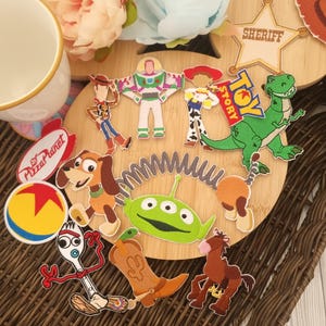 May include: A selection of embroidered patches featuring characters from the animated film "Toy Story." Patches include Woody, Buzz Lightyear, Jessie, Rex, and others. The patches are arranged on a wooden surface with floral accents.