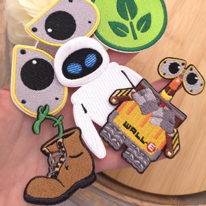 Puede incluir: Una colección de parches bordados con personajes y símbolos de la película WALL-E, incluyendo un robot blanco, un robot amarillo con el texto WALL-E, una bota marrón con una planta verde, dos piezas oculares grises y un círculo verde con hojas.