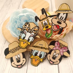 May include: A collection of embroidered patches depicting Disney characters Mickey, Minnie, Donald, Daisy, Goofy, and Pluto, each sporting safari hats. The patches are brightly coloured with black, yellow, and tan details, arranged on a wooden surface with floral accents.