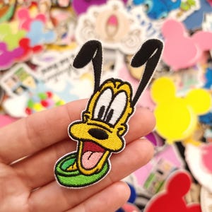 May include: Embroidered Pluto patch with a yellow body, black ears, and a green collar. The patch shows a smiling expression with a red tongue. The background is filled with various colourful patches and stickers, including Mickey Mouse shapes and other cartoon characters.