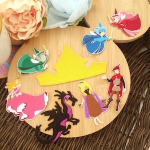 May include: A collection of colorful fabric patches featuring characters and elements from a fairy tale. The patches include a yellow crown, a black dragon, and various figures in pink, green, blue, and red attire, arranged on a wooden surface.