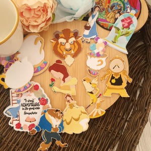 Può includere: Una collezione di toppe ricamate raffiguranti personaggi e scene del film d'animazione "La Bella e la Bestia". Include Belle, la Bestia e altri elementi iconici. Le toppe sono disposte su una superficie di legno con accenti floreali.