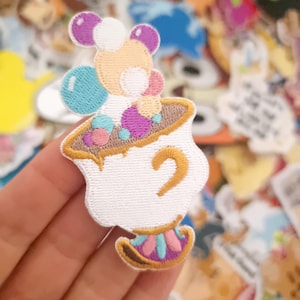 Puede incluir: Parche bordado con un personaje de dibujos animados en una taza de té. El personaje tiene un cuerpo naranja claro, orejas moradas y detalles azules. La taza de té es blanca con un asa dorada y una base colorida. El parche está rodeado de otros parches coloridos.