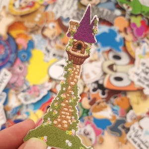 May include: Embroidered patch depicting a purple-roofed tower with a brown basket-like base and a green vine-covered stem. The tower is decorated with small flowers and sits on a green base. The patch is held against a backdrop of other colourful patches.