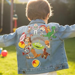 以下が含まれることがあります： 明るい青色のデニムジャケット。背中には、バズ・ライトイヤーやウッディなど、カラフルなトイ・ストーリーのキャラクターパッチが施されています。 太陽の下、屋外で子供が着用しており、背景にはブランコがあります。