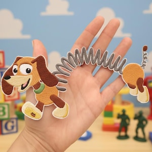 Puede incluir: Parche bordado de Slinky Dog de Toy Story. El parche presenta un perro marrón y canela con un cuerpo de resorte. El parche se sostiene en una mano contra un telón de fondo de bloques de juguete y un cielo azul.