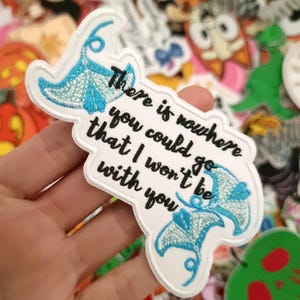Puede incluir: Parche bordado blanco con la cita "There is nowhere you could go that I won't be with you" en texto negro. El parche presenta dos diseños de manta rayas azules y un borde blanco. El fondo está lleno de varios parches de personajes de dibujos animados.