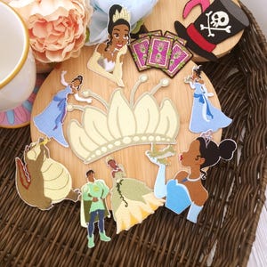 May include: A collection of embroidered patches featuring cartoon characters, including princesses, a frog, and a pirate hat with a skull and crossbones. The patches are arranged on a wooden surface within a woven basket, with floral accents.