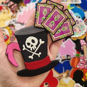 Pode incluir: Patches bordados com um chapéu alto preto com caveira e ossos cruzados, uma faixa vermelha e uma pena rosa. Também patches de cartas de baralho roxas com detalhes verdes e brancos. Para roupas ou acessórios.