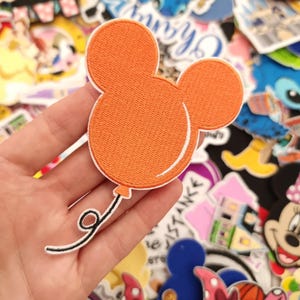 May include: An orange Mickey Mouse balloon patch with a white outline and a black and white string. The patch is held in a hand, with other colorful patches in the background.