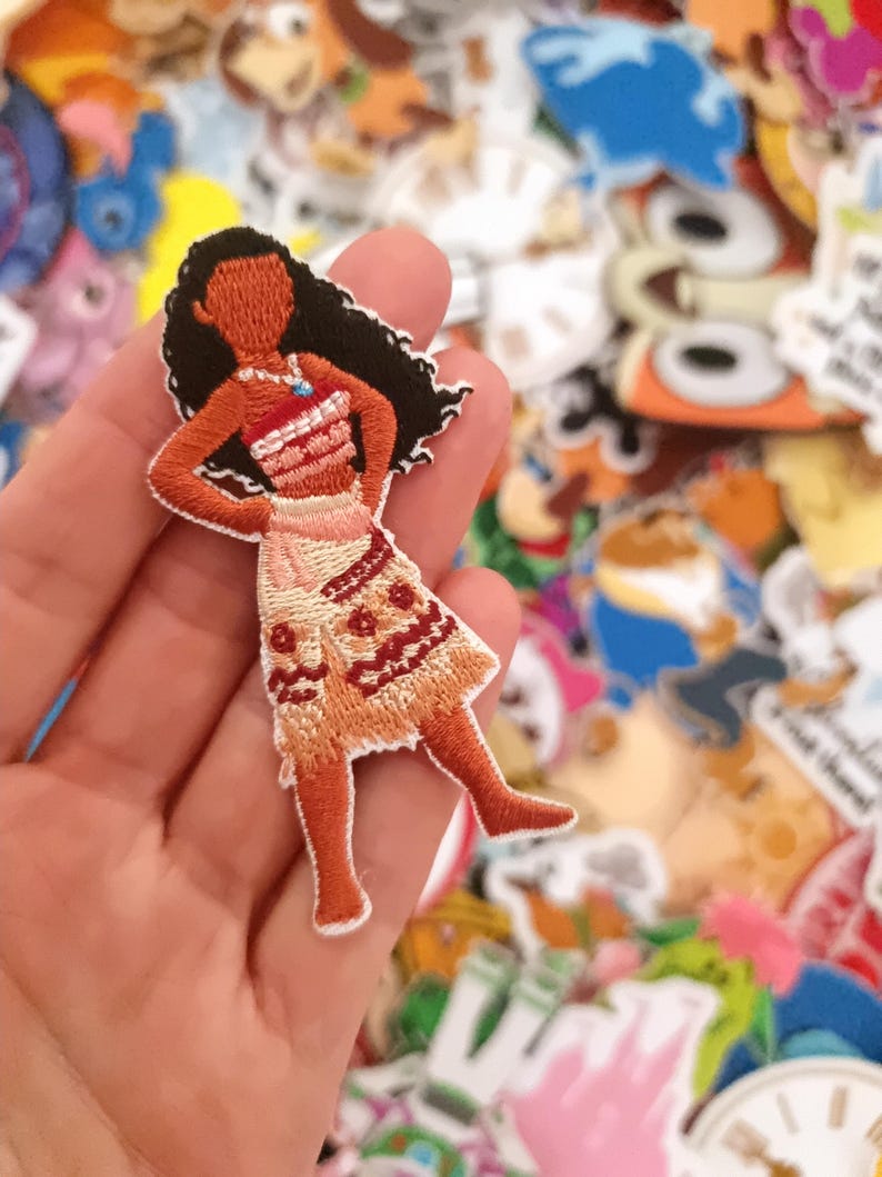 K&ouml;nnte beinhalten: Gestickter Aufn&auml;her mit einer Zeichentrickfigur mit langen schwarzen Haaren, die ein rot-wei&szlig;es Oberteil und einen gemusterten Rock tr&auml;gt. Der Aufn&auml;her wird in einer Hand gehalten, mit einem Hintergrund aus anderen bunten Aufn&auml;hern.