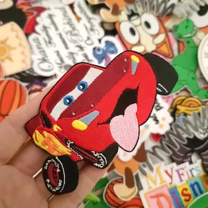 Puede incluir: Un parche bordado rojo de un coche de carreras de dibujos animados con una lengua rosa que sobresale. El parche tiene ruedas negras y detalles en amarillo y azul. El fondo está lleno de otros parches y pegatinas.