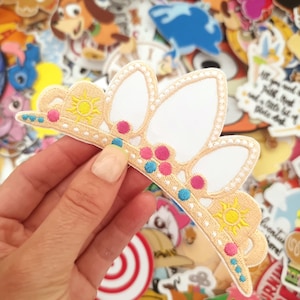 May include: A light tan and white embroidered crown patch with a sun design and pink and blue accents. The patch is surrounded by other colorful patches.