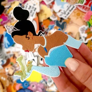 以下が含まれることがあります： 水色のドレスとティアラを着た漫画のプリンセスが描かれた刺繍パッチ。パッチは黒いお団子と茶色の肌をしています。パッチは手で持たれており、背景には他のパッチがあります。