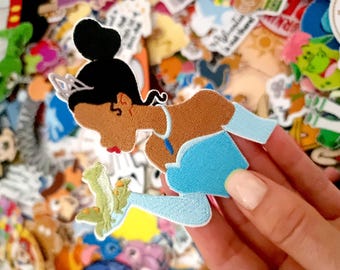 May include: An embroidered patch featuring a character with dark hair in a high bun, wearing a light blue off-the-shoulder dress and a silver tiara, holding a small green frog in one hand. The patch has a white border.