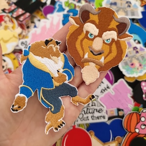 Può includere: Toppe ricamate raffiguranti la Bestia de La Bella e la Bestia. Una toppa mostra il volto della Bestia, l'altra la Bestia in abito blu. Le toppe sono marroni, blu e beige.