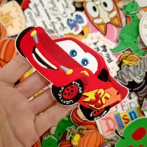 Può includere: Una toppa ricamata raffigurante un'auto da corsa rossa con occhi blu, una bocca larga e sorridente e un fulmine giallo sul lato con il numero 95 in rosso. L'auto ha pneumatici neri con scritte bianche e dettagli gialli.