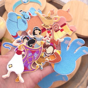 May include: A collection of embroidered fabric patches featuring characters and objects from the Aladdin story, including a blue genie, a monkey, a princess, a magic carpet, a golden lamp, and a palace, all rendered in vibrant, multi-colored thread.