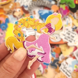 May include: Embroidered patch featuring a princess in a pink dress with yellow hair. The patch is held in a hand, with other colorful patches in the background. The princess has a pink bow and is in a dancing pose.