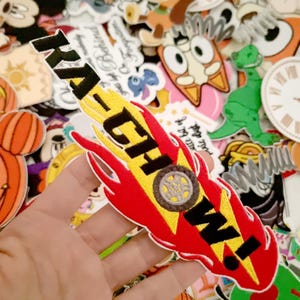 May include: A red, yellow, and black embroidered patch with the text "KACHOW!" in black. The patch has a flame design and a circular emblem in the center. The background features various cartoon character patches.