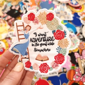 May include: Embroidered patch with a woman in a blue dress climbing a ladder, framed by a floral wreath. The text reads "I want ADVENTURE in the great wide Somewhere". The patch is white with red, pink, and beige details.