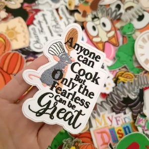 Può includere: Toppa ricamata con Remy di Ratatouille che tiene un cucchiaio di legno, con la citazione "Chiunque può cucinare, ma solo gli impavidi possono essere grandi". La toppa è bianca con dettagli neri e grigi, circondata da altre toppe a tema Disney.