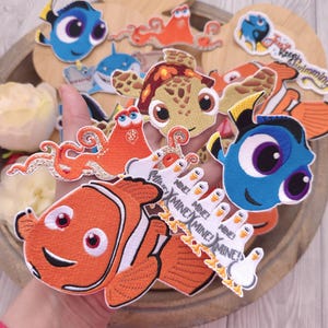 May include: A collection of embroidered fabric patches featuring various cartoon sea creatures and birds. The designs include a blue fish, an orange clownfish, a sea turtle, an orange octopus, and a group of white seagulls with the text MINE!