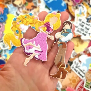 May include: Embroidered patches featuring a princess in a pink dress with long blonde hair and a prince in a blue and brown outfit. The patches are colorful and detailed, with a variety of other patches in the background.