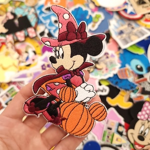 May include: Embroidered patch of a cartoon character in a witch costume, holding two orange pumpkins. The character wears a pink witch hat with white spots and a burgundy bow. The patch is surrounded by other colourful patches.