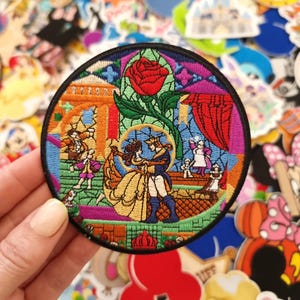 May include: A circular embroidered patch featuring a colorful scene of a couple dancing in a ballroom, surrounded by secondary characters and a large red rose at the top. The design is enclosed by a thick black border with intricate stitching.