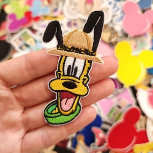 May include: Embroidered Pluto patch wearing a safari hat. The patch is yellow, black, and tan with a green collar. The patch is surrounded by other colourful patches with cartoon characters.