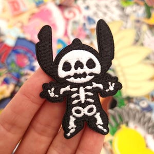 May include: A black and white embroidered patch depicting a cartoon character in a skeleton design. The patch has a black outline and white detailing, including the skull, rib cage, and limbs. The patch is held in a hand, with a colourful background of other patches.