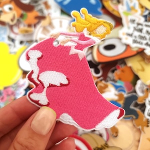 Könnte beinhalten: Gestickter Aufnäher mit einer Prinzessin in einem rosa Kleid mit weißer Bordüre und einer gelben Krone. Der Aufnäher ist von anderen bunten Aufnähern umgeben, was eine verspielte und skurrile Ästhetik erzeugt. Ideal zum Dekorieren von Kleidung oder Accessoires.