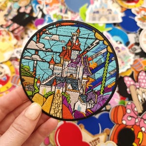 Op de afbeelding: Een ronde geborduurde patch met een kleurrijk kasteelontwerp met een blauwe lucht op de achtergrond. De patch heeft een zwarte rand en wordt door een hand vastgehouden. Andere kleurrijke patches en stickers zijn op de achtergrond.