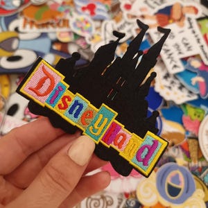 May include: A black embroidered patch featuring the silhouette of a castle with the word Disneyland stitched in colourful letters across the front. The text is displayed in a yellow rectangular border with pink, blue, and orange thread colours.