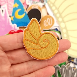 May include: Embroidered patch with a golden yellow seashell design. The patch has a detailed, textured appearance with a spiral pattern. Other colourful patches are visible in the background, suggesting a collection of decorative items.