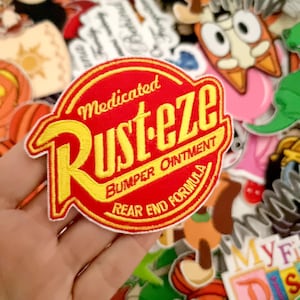 Pode incluir: Um patch bordado vermelho e amarelo com o texto "Medicated Rust-eze Bumper Ointment Rear End Formula". O patch é rodeado por outros patches e desenhos coloridos.