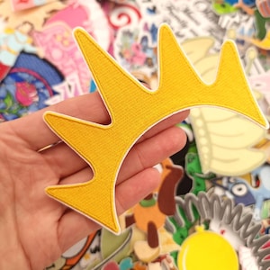 Puede incluir: Parche de corona bordada amarilla con ribete blanco, sostenido en una mano. El parche tiene una apariencia texturizada. Otros parches coloridos con temática de Disney están en el fondo, incluyendo una pancarta de "Disneyland".