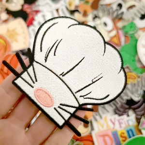 Può includere: Toppa ricamata raffigurante un cappello da chef bianco con dettagli neri e un ovale rosa. La toppa è circondata da altre toppe colorate, creando una scena giocosa. Perfetta per aggiungere a vestiti o accessori.