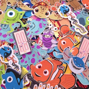 May include: A collection of embroidered patches featuring various animated characters, including a bright orange clownfish, a blue tang fish, a green one-eyed monster, a purple monster, and white seagulls with the text "MINE!" printed repeatedly.